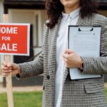 Real estate agent holding a for-sale sign in front of a property.