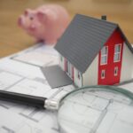 Miniature house, house plans, magnifying glass, and piggy bank emphasizing mistakes home buyers and sellers make.