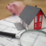 Magnifying glass, model house, and a piggy bank on top of blueprints